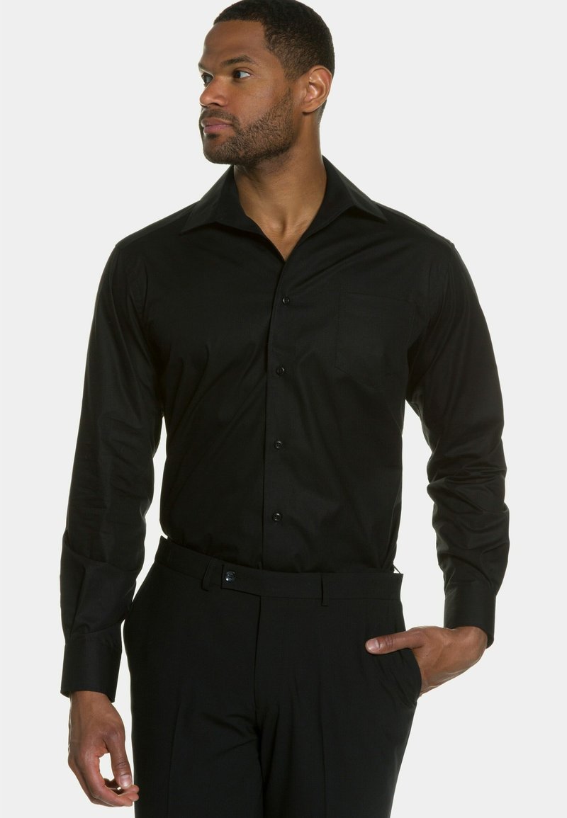 Camisa negra de manga larga con cuello abotonado, bolsillo frontal y textura suave. Se combina con pantalones negros, de corte ajustado y líneas limpias.