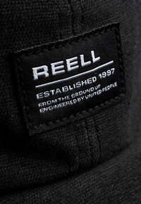 Schwarzes Stoffetikett mit weißem Text "REELL", gegründet 1997, sowie weiteren Details. Nahaufnahme zeigt strukturierten Stoff und Nähte.