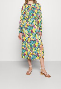 Blommig midiklänning med ett livligt mönster i blått, gult och grönt, långa ärmar, en sidoklyvning och en mjuk tygstruktur. Kompletterad med vita sandaler.