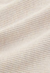 Detailní záběr na měkkou béžovou tkaninu s kroužkovou pleteninou, která ukazuje texturu a malé vyvýšené mřížkové vzory s jemnými záhyby.