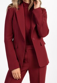 Blazer sur mesure bordeaux avec revers crantés, fermeture à un bouton et poches latérales. Porté sur un pull en maille côtelée bordeaux, mettant en valeur un look monochrome.