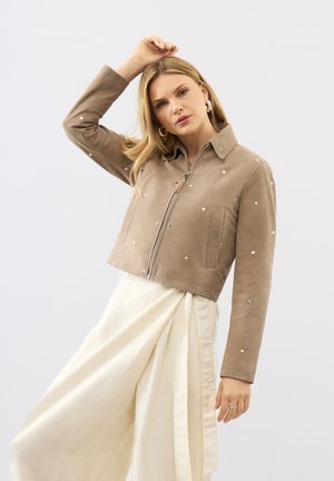 Vrouw poseert in een beige jas met rits en kleine studs en een vloeiende witte rok tegen een effen lichte achtergrond.