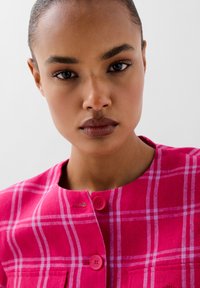 Chaqueta de cuadros en tonos rosas con cuello redondo, dos bolsillos y botones a juego. La tela tiene una apariencia texturizada, lo que realza la visibilidad del diseño en su conjunto.