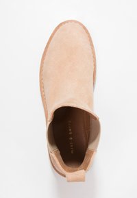 Bota de tobillo de ante beige con punta redonda, paneles laterales elásticos y un tirador de tela en la parte posterior. Superficies texturizadas y lisas.