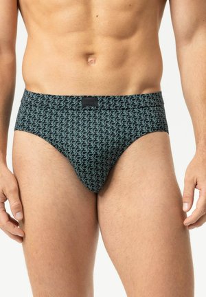 Männlicher Oberkörper mit dunklen Slips mit kleinem hellblauem Muster, stehend mit entspannten Armen an den Seiten vor einem schlichten Hintergrund.