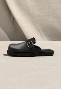 Sandale noire à bout fermé, à enfiler, avec une semelle épaisse et deux brides à boucles dorées sur le côté, posée sur un fond neutre.