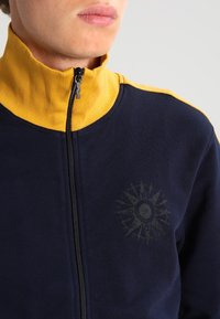 Marinblå zip-jacka med hög krage, gula detaljer och en kompassdesign tryckt i mörkgrått på bröstet. Materialet är en bomullsblandning.