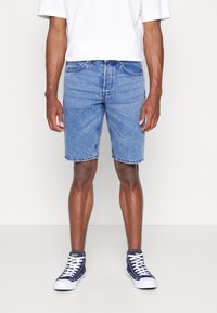 Pantalones cortos de mezclilla en azul claro. Diseño estándar de cinco bolsillos, corte recto, con dobladillo sin terminar. Combinados con zapatillas altas en azul marino y una camisa blanca.