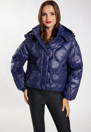 Frau trägt eine glänzende marineblaue gesteppte Steppjacke mit hohem Kragen und Kapuze, kombiniert mit schwarzen eng anliegenden Hosen.