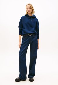 Camisola azul com capuz, bolso frontal e cordão, combinada com jeans escuros de cintura subida e sapatos pretos de plataforma. Roupa simples e casual.