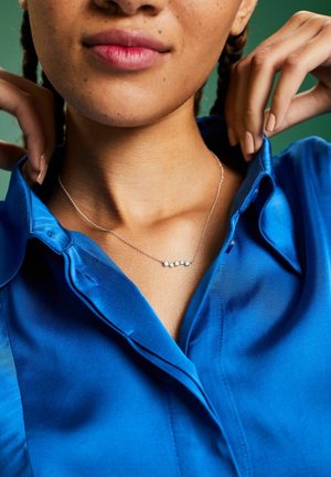 Collana d'argento con cinque piccoli accenti circolari, indossata sopra una camicia di satin blu con colletto. Presenta una texture liscia e una lucentezza brillante.