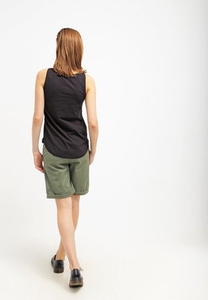 Femme aux cheveux mi-longs marchant de dos, portant un haut sans manches noir, un short vert olive et des chaussures noires sur fond blanc.