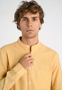 Camicia gialla a maniche lunghe con colletto alla cinese, taschino sul petto e tessuto strutturato. I bottoni sono bianchi e rotondi.