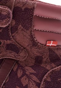 Bordeaux suède sneaker detail met bloemtextuur, gestikte accenten, een soepel leren paneel en een klein rood label met een wit kruispatroon.