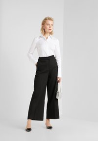 Chemise blanche à boutons, pantalons larges noirs taille haute, mules noires avec détail de nœud, et un petit sac à main en chaîne argentée.