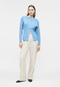 Frau steht und trägt eine hellblaue, geknöpfte Strickjacke, lockere, cremefarbene Hosen und schwarze Ballerinas vor einem schlichten weißen Hintergrund.