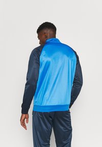 Giacca a track blu e navy con zip frontale, polsini a costine e una consistenza morbida e liscia, caratterizzata da contrasti di colore.