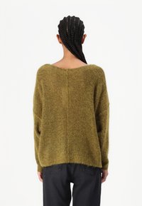 Olivovo zelený pletený sveter so širokým výstrihom, spadnutými ramenami a voľným strihom. Mäkká textúra s mierne chlpatejším povrchom, v kombinácii s tmavými džínami.