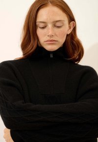 Zwart gebreid sweater met een hoge kraag en een kwart rits. Heeft een geribbelde textuur en kabelbrei-details op de mouwen.