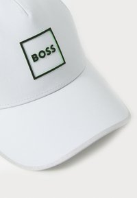 Bijela kapa od tkanine s zakrivljenim vizirjem. Ima crni kvadratni logotip s zelenim detaljima i tekstom "BOSS" u središtu.