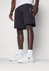 Svarta bomullsshorts med elastisk midja och sidofickor, med vit "Champion"-logga broderad. Försedda med vita sneakers och strumpor.
