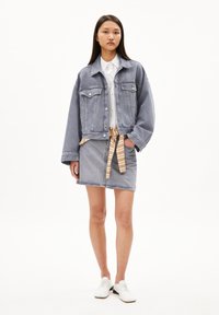 Chaqueta de denim de color azul claro con dos bolsillos en el pecho, combinada con una falda de denim azul claro que presenta un cinturón de rayas marrón. Unas zapatillas blancas completan el atuendo.