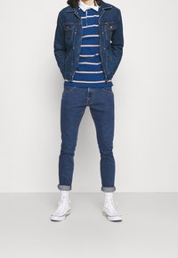 Giubbotto di denim blu scuro, polo a righe blu e arancione sotto, abbinato a jeans tapered blu scuro e sneakers alte bianche.