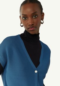 Niebieski dziany kardigan z dekoltem w serek, z zapięciem na jeden guzik i krótkimi rękawami. Noszony na czarnym golfie.