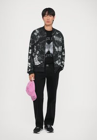 Giubbotto bomber nero con stampa floreale e di farfalle, indossato su una t-shirt grafica nera. Pantaloni neri e cappellino rosa in mano, sneakers nere.
