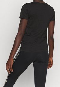Persona vestida con una camiseta de manga corta negra y leggings negros con una franja de logotipo multicolor en el costado, de pie contra un fondo liso.