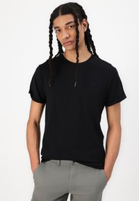 Black short-sleeve t-shirt with a round neck and rolled sleeves, made from smooth fabric. Subtle logo on the chest, worn with light gray pants.