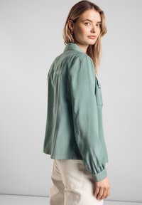 Světlá zelená, hedvábná blůza s volným střihem a manžetami na knoflíky. Obsahuje zadní yoke a dvě přední kapsy. Kalhoty v neutrálních tónech.