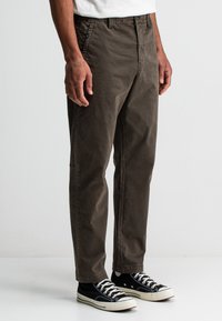 Bruine katoenen broeken met een relaxed fit, met een ritssluiting, zijzakken en een versleten textuur. Gecombineerd met zwarte canvas sneakers.