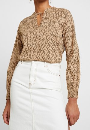 Woman wearing beige long-sleeve blouse with small black leaf pattern tucked into high-waisted white skirt with visible seam and pocket details.