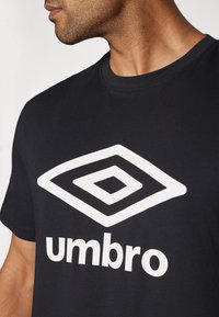 Sort bomulds T-shirt med et stort hvidt Umbro-logo. Designet inkluderer en diamantformet grafik og rund halsudskæring.