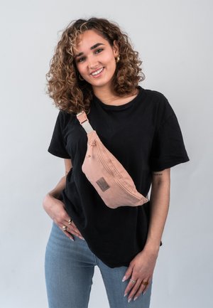 Pink fabric fanny pack with a zippered closure and a textured patch, worn crossbody over a black t-shirt, paired with light blue jeans.