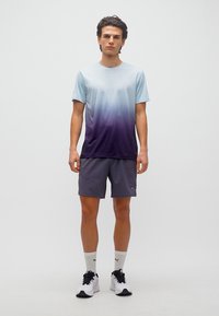 Νεαρός άνδρας που στέκεται, φοράει μπλε-μοβ degradé κοντομάνικο πουκάμισο, σκούρα γκρι σορτς, λευκές κάλτσες Puma και λευκά αθλητικά παπούτσια.