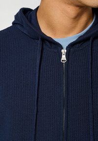 Le sweat à capuche zippé marine présente une texture côtelée, une capuche réglable et une fermeture éclair argentée. Une couche bleu clair est également visible en dessous.
