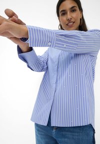 Blå og hvidstribet skjorte med lange ærmer, der har små dekorationer og en lige kant, båret med denim.