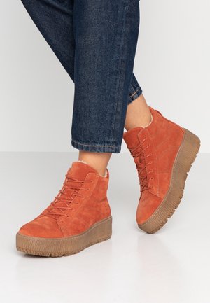 Personne portant des baskets montantes à plateforme en daim orange avec des semelles marron et un jean court bleu foncé sur fond blanc.