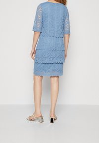 Robe en dentelle bleue à manches courtes, avec un ourlet à volants superposés, mettant en valeur des motifs floraux. Portée avec des sandales à talons blocs argentées.