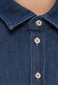 Denim shirt with a dark blue color, pointed collar, and visible stitching. Features metal buttons with a central hole. Close-up view.