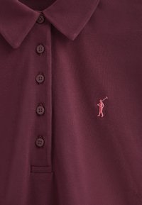 Burgundy pikétröja med krage, fem knappar och en liten rosa golfare-logotyp broderad på bröstet. Texturerad tyg.