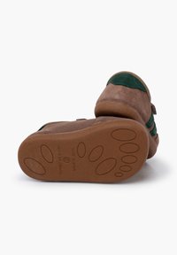 Chaussons en suède marron avec des accents en textile vert, bout arrondi et semelle en caoutchouc plate avec un motif antidérapant. Le texte sur la semelle indique l'origine.