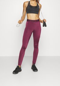 Bordowe legginsy z wysokim stanem, z czarnym logo, gładką fakturą i bocznymi kieszeniami. W parze z czarnym stanikiem sportowym i trampkami.