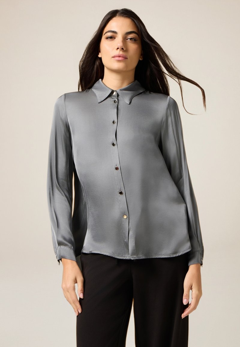 Blusa in satin grigia con colletto alla francese, maniche lunghe e chiusura a bottoni. Vestibilità ampia, con una texture liscia e una leggera lucentezza.