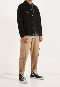 Svart jeansjacka med silverknappar, grå texturerad skjorta under, beige cargo-byxor med fickor och svarta högstövlar.