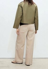 Personne portant une veste courte vert olive, un pantalon large beige et des sandales noires, vue de dos devant un mur blanc uni.
