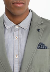 Mężczyzna w jasno szarym koszuli w prążki z guzikiem oraz stonowanej zielonej marynarce, z granatową poszetką i przypinką w kropki na klapie.