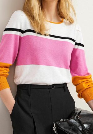 Femme portant un pull à manches longues avec des blocs de couleur blanc, rose, noir et orange, associé à un pantalon noir taille haute et tenant un sac noir.